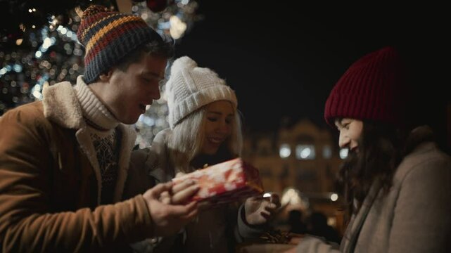 Friends presenting gift to each other outdoor at decorated illuminated background. Happy friends celebrate Christmas New Year Xmas Eve night together smiling and hug. Young people have joyful time
