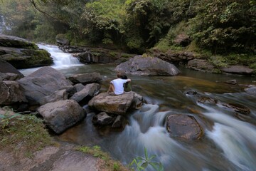 Person sitting by a river in a forest with a waterfall.