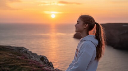 Fototapeta premium Stock minimalist photograph of a thoughtful teenage girl smiling gently while watching a sunset, standing on a cliff edge with ample open space and the soft, warm glow of the setting sun