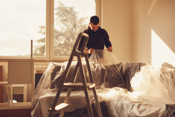 Man covering home couch with plastic sheets for protection against dust during renovation.