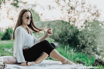 Young woman in sunglasses sitting on a blanket outdoors, enjoying a sunny day in a park.