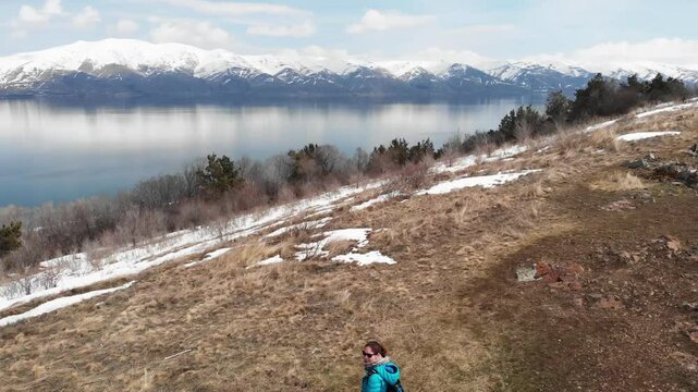 Aerial footage of a woman traveler admiring Lake Sevan in Armenia with calm blue water and snow mountains in the background The scene captures peaceful exploration and natural beauty of the landscape