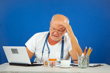 An elderly man, a doctor, in his office, at the table. Blue background.