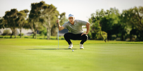 Fototapeta premium Focused male golfer crouching near a golf ball on the putting green on a picturesque golf course