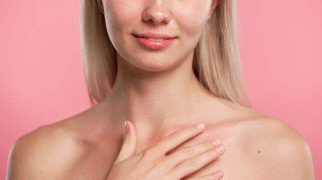 Young blonde woman posing at camera, applies body lotion on collarbone area, isolated on pink studio background close up shot. Beauty skin care concept