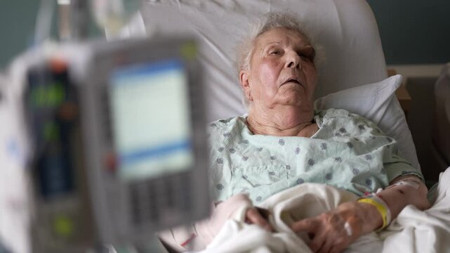 An elderly woman lies in a hospital bed, wearing a hospital gown and receiving IV fluids. A medical device is visible in the foreground, suggesting ongoing medical care.