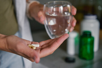 Woman holds two pills in one hand and glass of water in other. Person takes dietary supplements and vitamins. Daily medical procedure to maintain health