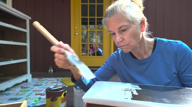 A woman in a blue shirt paints the top edge of a wooden drawer white with a paintbrush. The drawer is open and is sitting on top of another drawer. There is a paint can, a newspaper, and a pair of gla