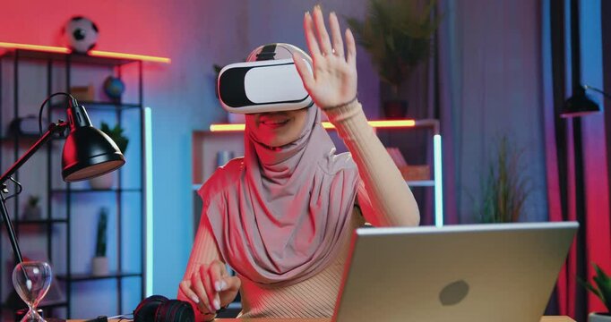 Woman in hijab working on imaginary screen in augmented reality goggles sitting at the desk at home in the evening