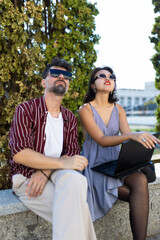 Young adults sitting outdoors using laptop while enjoying sunny weather in urban setting