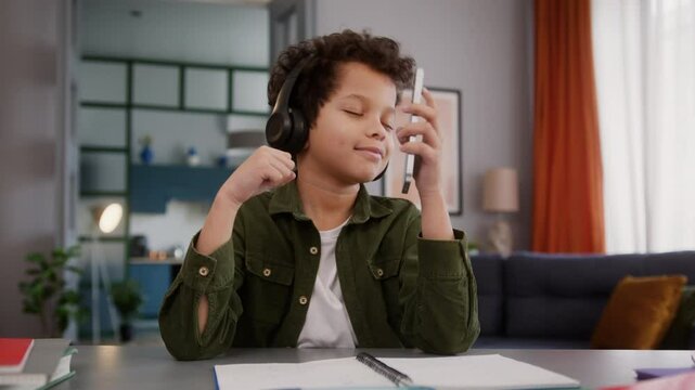 Funny African American son kid child boy pupil student schoolboy at home studying learning study learn pause break listening music in headphones listen audio song dancing enjoy dance holding phone