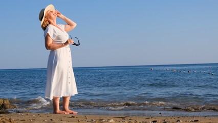 woman in a straw hat and white dress walks along the seashore puts on sunglasses and admires the blue sea on a sunny summer day, enjoying freedom and relaxation. The concept of a senior citizen