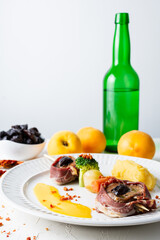Top view of sirloin skewer with broccoli, potato and sauce, on a plate and white background, with a bottle of cider and peaches, vertical, with copy space