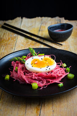Top view of rice noodles with beetroot, green asparagus and boiled egg, on black plate on wooden table with sauce and chopsticks, vertical, with copy space