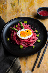 Overhead view of rice noodles with beetroot, green asparagus and boiled egg, on a black plate on a wooden table, vertical, with copy space