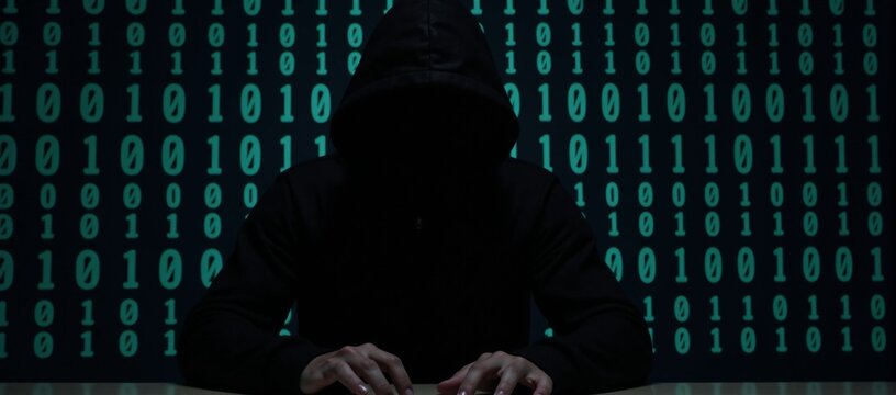 A hooded criminal silhouetted while hacking on binary code background cybercrime concept