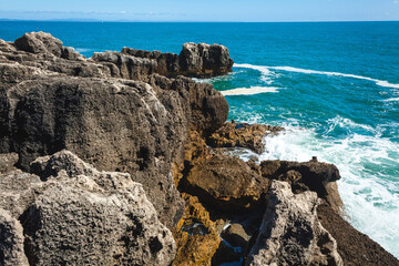 A peaceful ocean scene with shimmering blue water stretching to the horizon, rocky formations in the foreground, and a lone rock emerging from the waves