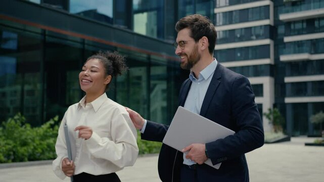 Two happy smiling friendly talking multiracial colleagues business co-workers walk in city African American woman businesswoman female girl talk Caucasian man businessman guy male hold laptop folder