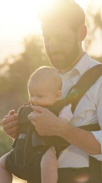 Cute attractive young father with a little newborn baby in a slings carrier. Use phone to take selfies on street near lake in park at sunlight. Ergo backpack. Parents. Gadget. Smartphone. Cell. Slow