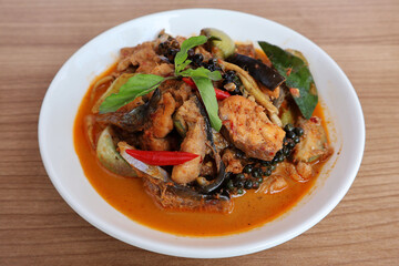 Spicy stir-fried giant catfish Red liquid, fish pieces, fresh pepper, brinjal eggplant, basil leaves in a cream-colored plate on a wooden table.

