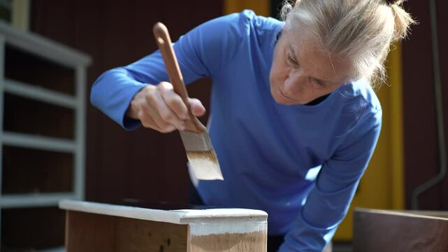 A woman wearing a blue shirt paints the top edge of a wooden dresser drawer with a white paint brush. The dresser is partially painted white. 