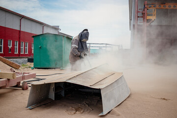 Worker sandblasting a metal structure to eliminate rust at an industrial plant during daylight