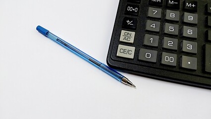 a blue pen lying on a white surface next to a black calculator. The pen has a blue cap and a metallic tip. The calculator is partially visible, showing a section of its keypad with large buttons.