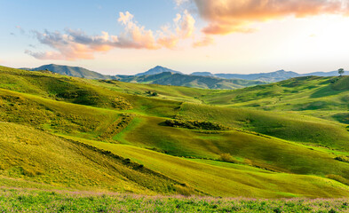 green spring season landscape of beautiful grassland hills in countryside with villages and mountains under scenic cloudy sky