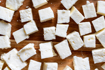 Block of raw cottage cheese or Indian paneer cut into small cubes on wooden chopping board. Top view.