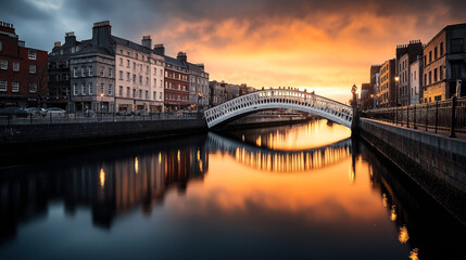 Naklejka premium Historical pedestrian bridge over a calm river at sunset, flanked by old stone buildings along the waterfront, reflecting vibrant orange and blue hues in the water surface.