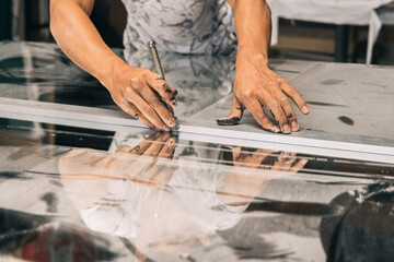 A close up of hands holding a glass cutter, scoring a piece of glass along measured lines in a...