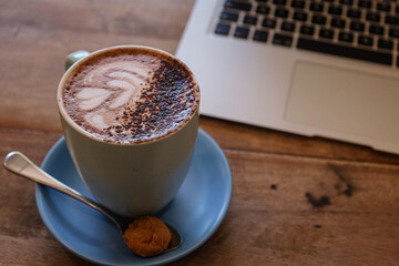 Cup of coffee on a table with laptop in the background. Hot chocolate in a mug. Latte art with a heart and chocolate. Hot drinks for breakfast at the cafe. Working at the cafe. Laptop and coffee.