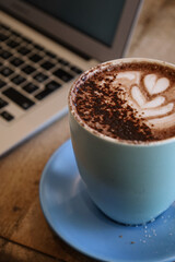 Cup of coffee on a wooden table with laptop in the background. Working at the cafe with a hot chocolate. Latte art in a blue mug. Hot drinks for the breakfast at the cafe. Portrait picture coffee.
