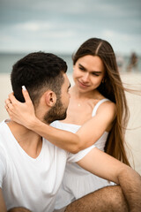 Young girl hugs her husband by the neck and looks into his eyes smiling while sitting on the sand at the beach