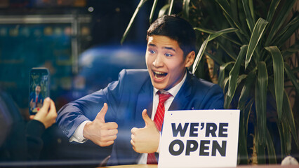 Young smiling Asian man in blue suit taking photo with we are open sign and thumbs-up, showing excitement about opening new venture. Concept of new business, startup, achievement, progress