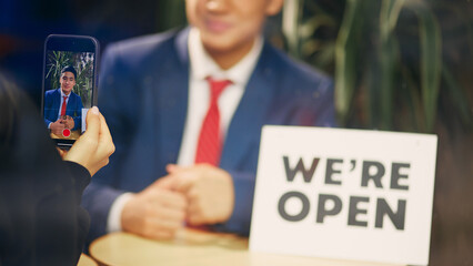 Behind glass winding, someone taking a photo of businessman in blue suit sitting with we are open sign. Social media post, news. Concept of new business, startup, achievement, progress