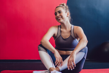 Positive hipster girl dressed in stylish tracksuit smiling while taking rest on red carpet for pilates training, happy female athlete enjoying time for crossfit in sport hall feeling carefree