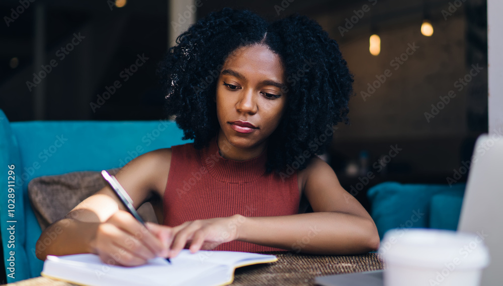 Poster dark skinned hipster girl writing information in knowledge notepad while sitting at table with moder