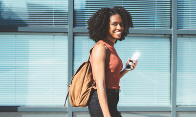 Full length portrait of cheerful African American shopper with paper bags in hands smiling at camera while strolling to next store with good offers of sales on Black Friday, happy woman in city