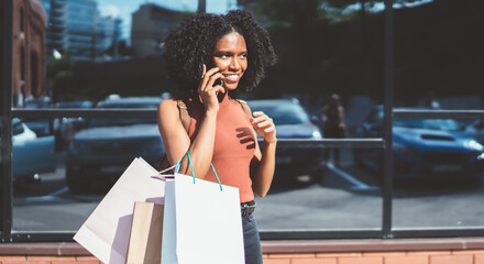 Happy black female teenager with paper bags calling to friend for discussing Cyber Monday discounts using roaming connection on cellphone, positive woman in casual wear smiling from funny joke