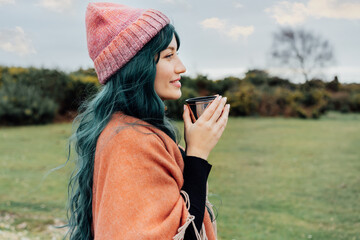 Portrait of smiling Woman in hat wrapped in Shawl Poncho drinking hot drink from thermos cup enjoying autumn landscape view. Feeling harmony, relax, personal fulfilment. Local travel lifestyle
