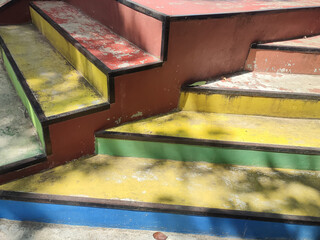 A set of colorful, weathered stairs with shades of red, yellow, green, and blue under sunlight, creating an artistic and vibrant scene.