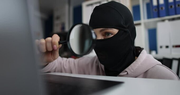 Robber wearing a mask and searching for information using magnifying glass on computer in office. Cybercrime and internet scammer concept