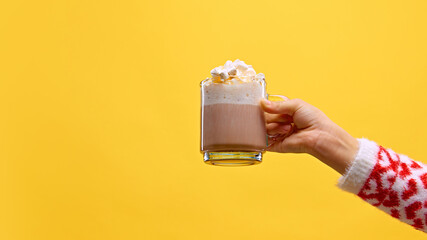 Hand in cozy sweater holds glass mug of hot chocolate topped with fluffy marshmallows against warm...