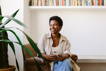 Thoughtful woman smiling on sofa, holding mobile phone and looking away