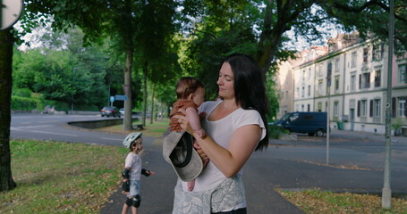 Mother holding baby on a quiet urban street, smiling at her child, surrounded by trees and greenery, with a young child on a skateboard nearby, peaceful family moment
