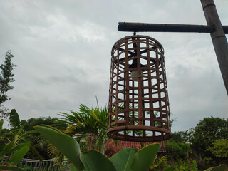 A rustic bamboo lampshade hangs from a wooden beam against a backdrop of lush greenery and a cloudy...