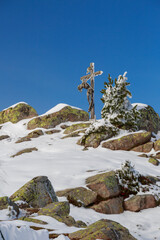 Holzkreuz auf schneebedeckten Felsen, davor ein klarer blauer Himmel in winterlicher Landschaft