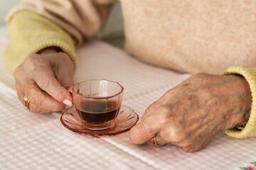 Elderly woman's hands holding a small pink cup of coffee; pastel colors - yellow and pink