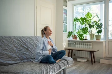 Fototapete Calm woman sitting in lotus pose deep breathing during meditation at home. Self-help of middle aged female stress relief in daily routine reset mind with eyes closed, placing hands on chest, abdomen. © DimaBerlin